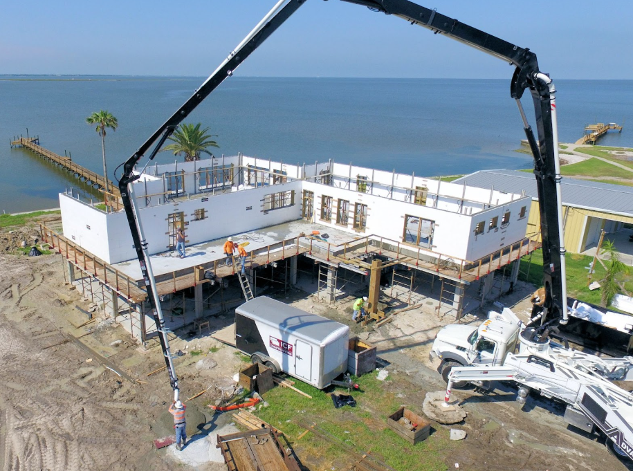 ICF home under construction by the water, with workers and concrete pumps on site.